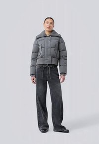 Woman wearing a gray padded jacket with large collar and black wide-leg jeans, standing against a plain light gray background.