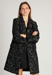 Mujer con cabello largo castaño que lleva un abrigo negro con estampado floral sobre un vestido negro texturizado, de pie con los brazos cruzados frente a un fondo sencillo.