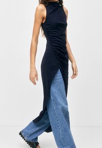 Vestido sin mangas de color azul marino con detalles fruncidos, cuello alto y abertura lateral, combinado con pantalones de mezclilla de lavado claro de pierna ancha y calzado negro grueso.