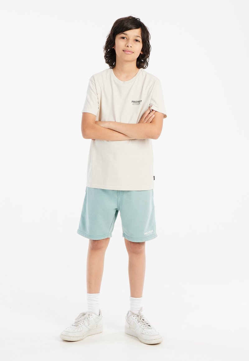 Crème T-shirt met een zwart logo, gecombineerd met mintgroene shorts met een logo en witte sneakers met grijze accenten. Witte achtergrond.