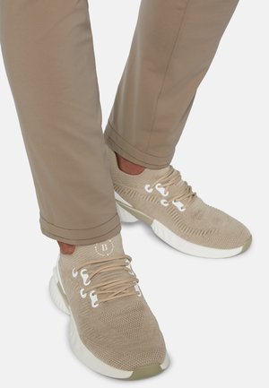 Beige sportsko med mesh-overdel, tekstureret strikdesign, hvid sål og subtilt logo på tungen, parret med lysebrune tapered bukser.