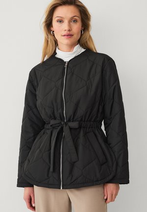 Kobieta ubrana w czarną pikowaną kurtkę z przednim zamkiem błyskawicznym, paskiem do wiązania i kieszeniami, w połączeniu z beżowymi spodniami i białym golfem.