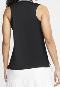 Žena viděná zadu, oblečená v černém bezrukávovém polokošili a bílých sukni, stojící před jednoduše bílým pozadím.