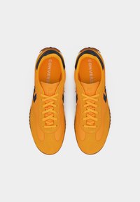 Ζευγάρι φωτεινά πορτοκαλί αθλητικά παπούτσια Converse με μαύρες λεπτομέρειες και λευκό κείμενο "Converse" στις εσωτερικές σόλες, εμφανιζόμενα από εφαπτόμενη όψη σε λευκό φόντο.