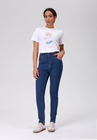 Camiseta gráfica branca com ilustração, jeans denim azuis de cintura alta e ténis cinzentos. Ajuste justo com uma textura suave.
