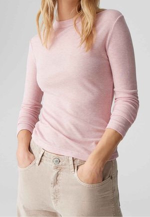 Femme portant une chemise rose clair à manches longues et un pantalon beige, les mains dans les poches avant, sur un fond uni.