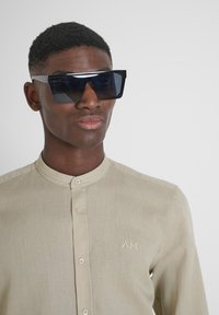 Occhiali da sole neri oversize con lenti angolari, indossati con una camicia col beige caratterizzata da un design con bottoni e dettagli ricamati.