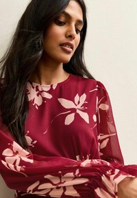 Vrouw met lang donker haar, gekleed in een doorschijnende burgundy top met lange mouwen en lichtroze bloemmotieven, kijkt omlaag met een neutrale uitdrukking.