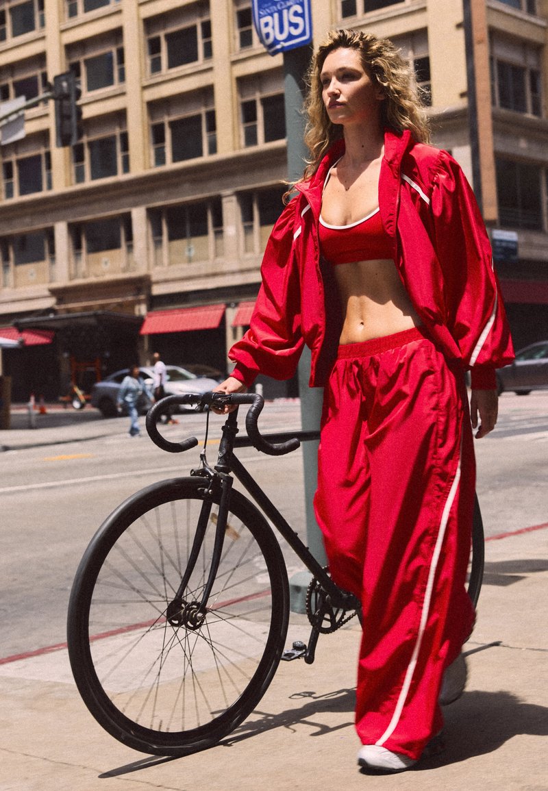 Femme en tenue de sport rouge marchant avec un vélo noir sur le trottoir en ville près d'un arrêt de bus en journée.