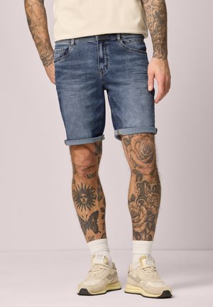Mann trägt hochgekrempelte blaue Jeansshorts, weiße Turnschuhe und Socken, zeigt tätowierte Beine und Arme vor neutralem Hintergrund.