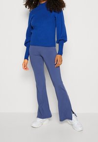 Blå stickad tröja med puffärmar som kombineras med lila utsvängda byxor med sidofickor och vita sneakers. Texturerad, åtsittande design.