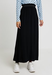 Gonna maxi nera con vita elasticizzata, realizzata in un tessuto morbido. Presenta una texture liscia e arriva fino alle caviglie. Indossata con sneakers bianche.