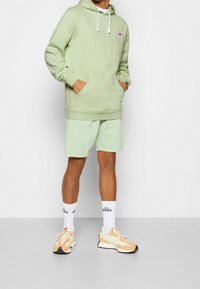 Felpa verde menta con tasca a marsupio, abbinata a pantaloni taglia corta dello stesso colore. Sneakers bianche con dettagli arancioni e calze con logo completano l'outfit.