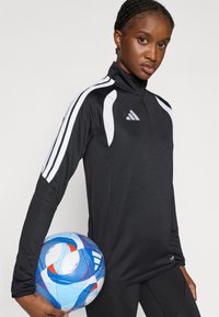 Μαύρο αθλητικό τοπ μακρυμάνικο με λευκές ρίγες και φερμουάρ στον γιακά, φορεμένο με μαύρα κολάν. Μια πολύχρωμη μπάλα ποδοσφαίρου κρατείται κάτω από το ένα χέρι.