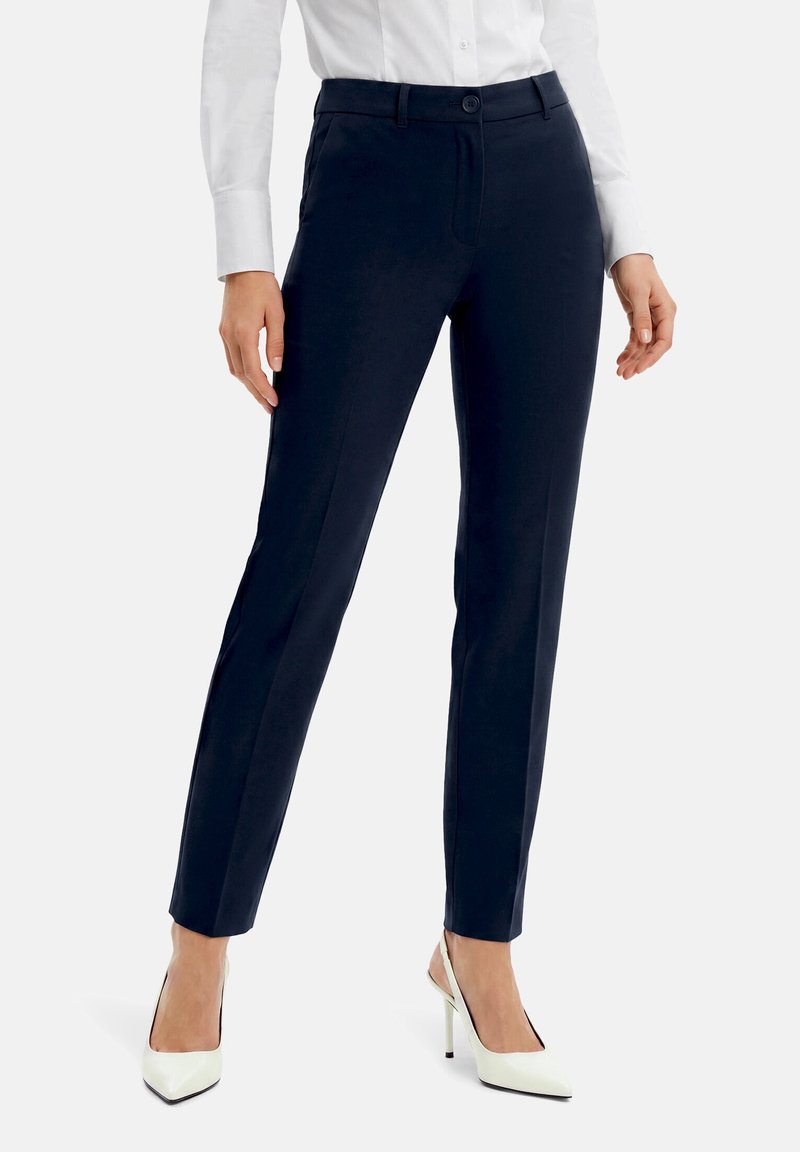 Vrouw die marineblauwe slim-fit pantalon, wit gebutton-down overhemd en witte hakken draagt, staand tegen een effen achtergrond.