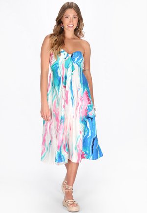 Jeune femme en robe bustier mi-longue avec un motif tourbillonnant bleu, rose et blanc, portant des sandales et des boucles d'oreilles colorées, souriante.