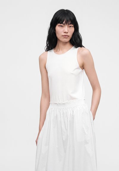 Femme aux longs cheveux noirs portant un haut blanc sans manches et une jupe blanche taille haute avec des poches sur un fond uni.