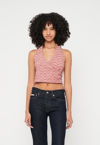 Kvinna med lockigt hår iförd en ärmlös rosa stickad halterneck crop top och mörkblå högmidjade jeans framför en enfärgad bakgrund.