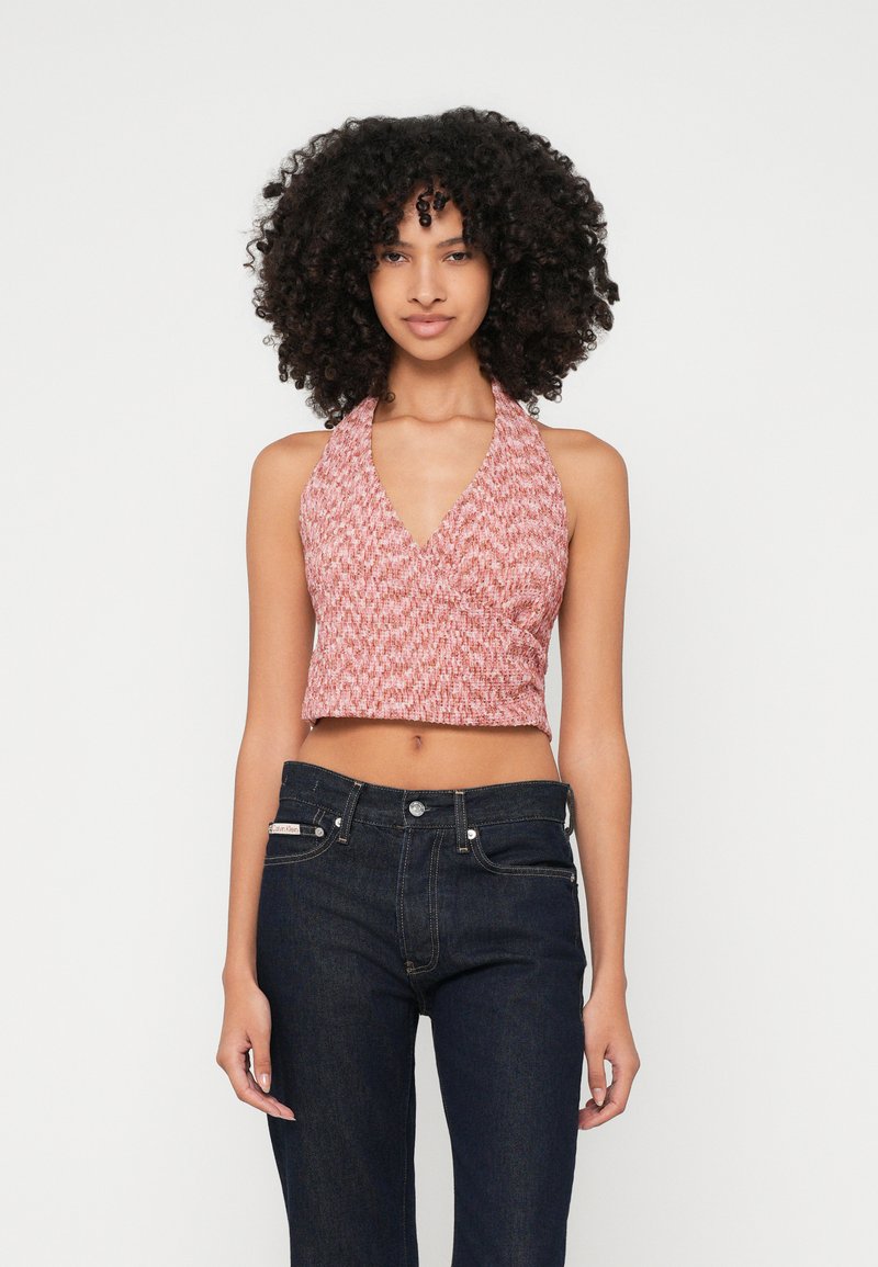 Kvinna med lockigt hår iförd en ärmlös rosa stickad halterneck crop top och mörkblå högmidjade jeans framför en enfärgad bakgrund.