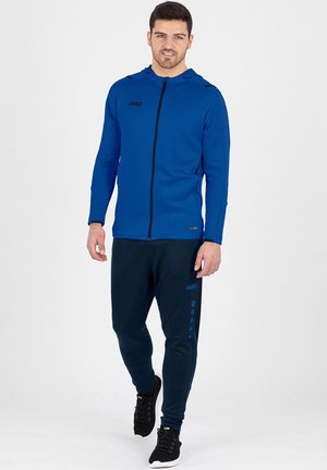 Mann trägt blauen Kapuzenpullover mit Reißverschluss und marineblaue Jogginghose, dazu schwarze Sneakers, steht und schaut nach vorne vor weißem Hintergrund.