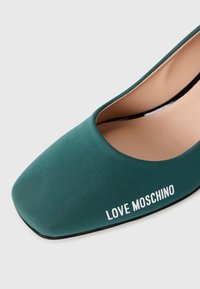 Žali audiniai bateliai su kvadratiniu priekiniu gaminiu. Išorėje yra kontrastingas baltas "LOVE MOSCHINO" logotipas ir lygus vidinis pamušalas.