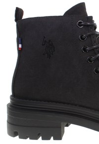 Black suede ankle boot featuring lace-up design, textured black rubber sole, and embossed logo detail. Accented with a tri-color tag.