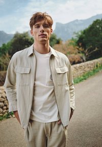 Lichtbeige jas met twee borstzakken over een witte t-shirt met een zakontwerp. Beige broek maakt de outfit compleet, buitenopname.