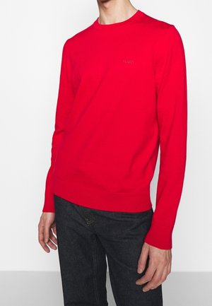 Homme portant un pull rouge vif à manches longues avec le logo "HUGO" sur la poitrine et un jean foncé, debout devant un fond blanc uni.