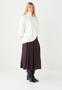 Weißer Rollkragenpullover mit Seitennähendetail, kombiniert mit einem braunen plissierten Midirock und schwarzen kniehohen spitzen Stiefeln.
