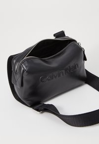 Černá kožená taška se zipovým uzávěrem, s embosovaným logem "Calvin Klein" a odnímatelným černým textilním popruhem. Hladký povrch a strukturovaný tvar.