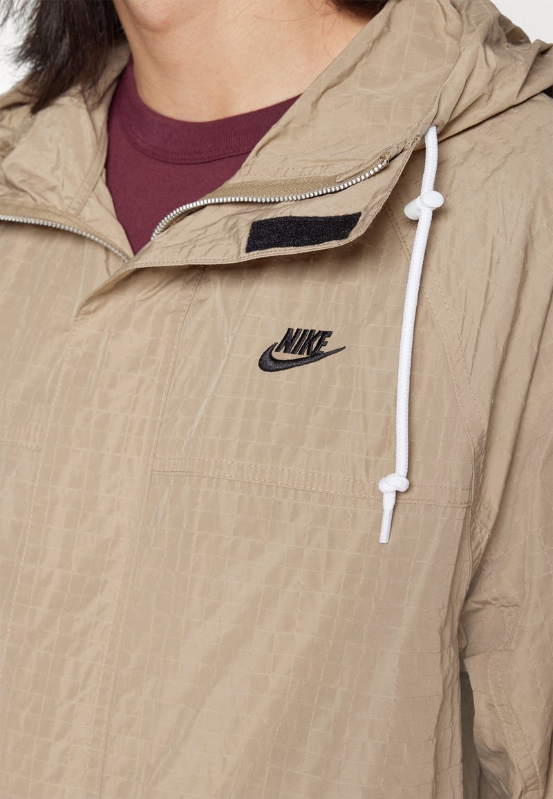 Chaqueta Nike beige con una superficie texturizada, que cuenta con un cierre en la parte delantera, cordón blanco y logo negro en el pecho.