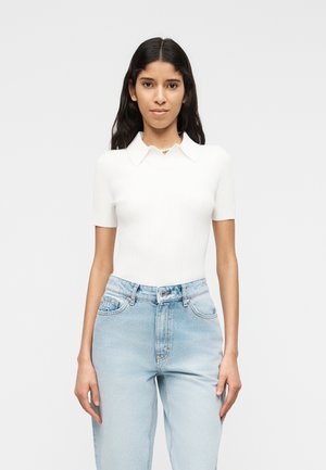 Młoda kobieta o długich ciemnych włosach, ubrana w biały krótki rękaw z kołnierzykiem i jasnoniebieskie dżinsy z wysokim stanem, stojąca na jednolitym tle.