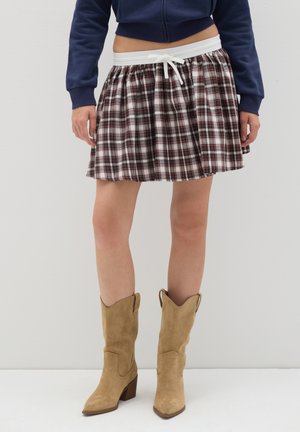 Jupe à carreaux en tissu marron et blanc avec un design froncé, dotée d'une taille élastique blanche et d'un cordon de serrage. Portée avec des bottes de cowboy beige.
