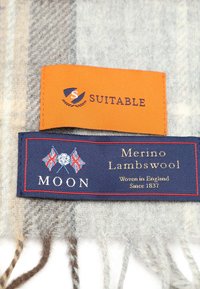 Etikett eines Merinowollschals aus Lammwolle mit marineblauem Hintergrund, rotem Rand und Emblemen in Weiß und Blau. Enthält ein orangefarbenes Etikett mit der Aufschrift "Geeignet."