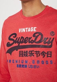 Mężczyzna w czerwonym swetrze z napisem "Vintage SuperDry" i japońskimi znakami nadrukowanymi na przodzie.