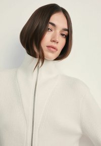 Sweter z wysokim kołnierzem, zapinany na zamek, w białym kolorze. Posiada pionową, żebrowaną strukturę i wzór w kształcie chevronu z przodu. Miękka, lekką tkanina.