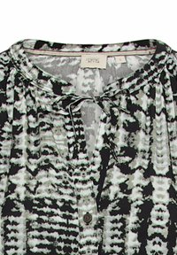 Blouse à motifs noir et blanc avec un col rond, un détail de nœud, une fermeture par bouton et un tissu léger et texturé.