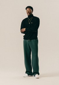 Άνδρας που στέκεται με τα χέρια σταυρωμένα, φορώντας γυαλιά, μαύρο πουλόβερ, χρυσά κολιέ, ρολόι, σκούρα πράσινα παντελόνια και λευκά αθλητικά παπούτσια σε απλό φόντο.