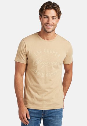 Homme aux cheveux châtain clair portant un t-shirt beige Lee Cooper et un jean bleu, souriant sur un fond blanc.