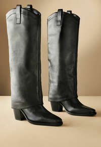 Botas de cuero negro hasta la rodilla con una textura suave, punta cuadrada y tacón grueso. Presentan un diseño de solapa y bucles en la parte superior para estilizar.