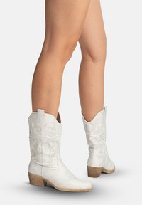 Benedenbenen met witte leren cowboylaarzen tot halverwege de kuit, met bloemrijke borduursels en gestapelde houten hakken, staand op een witte ondergrond.