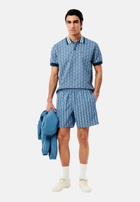 Camisa polo azul con estampado, de manga corta y cuello, combinada con pantalones cortos a juego. Una chaqueta azul claro y zapatillas de crema completan el look.