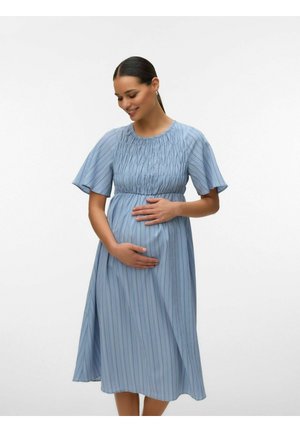 Gravid kvinne i blå stripete kjole med korte ermer, som holder magen forsiktig og smiler mot en ensfarget hvit bakgrunn.