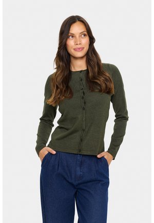 Cardigan long à manches longues vert olive avec une finition texturée, présentant un col rond et six boutons sur le devant, associé à un pantalon en denim large.