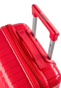 Valigia rigida rossa con superficie texturizzata, dotata di manico in alluminio retrattile e chiusura a zip.