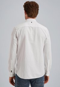 Camisa blanca de lino con cuello tipo botón, mangas largas y puños abotonados. Presenta una textura sutil y un pequeño logotipo en la parte posterior.