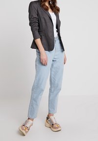 Veste grise à épaules structurées, coupe à simple boutonnage avec trois boutons, associée à un jean taille haute bleu clair et des baskets multicolores.