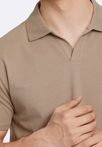 Muž v béžovém polo tričku s krátkým rukávem, který si pravou rukou upravuje nebo se dotýká oblasti límce.