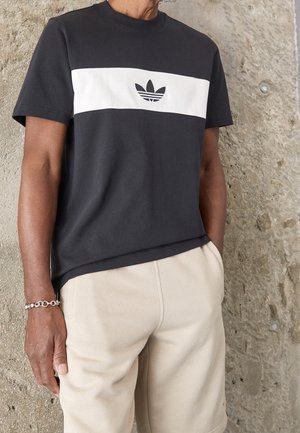 Homme portant un t-shirt Adidas noir et blanc et un short beige, debout contre un mur de pierre texturé.