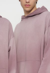 Νεαρό άτομο με ανοιχτό μωβ φούτερ με κουκούλα, φαίνεται από τον ώμο μέχρι το πιγούνι σε απλό ανοιχτό φόντο.